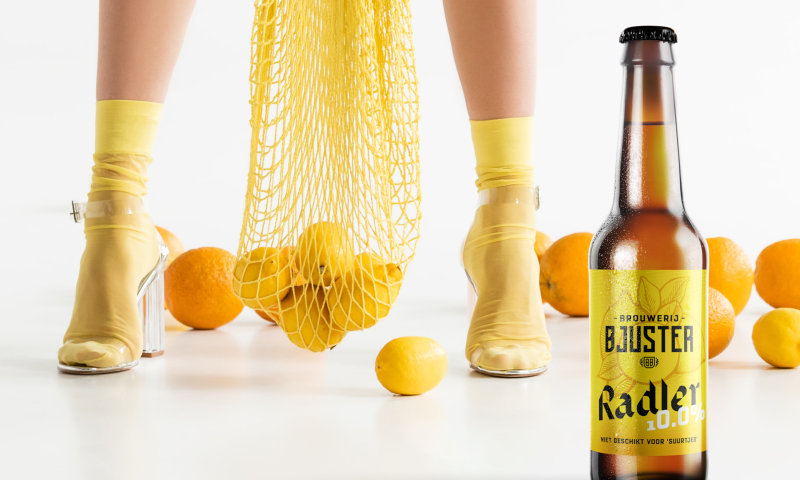 Bjuster Radler 10% flesje met dame erachter met een netje citroenen en sinaasappels